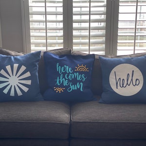K&ouml;nnte beinhalten: Drei marineblaue Zierkissen mit wei&szlig;en Designs. Eines zeigt ein Sonnenstrahl-Design, ein anderes den Text "here comes the sun" mit Regenbogen und Sonne, und das dritte sagt "hello" in einem wei&szlig;en Kreis.