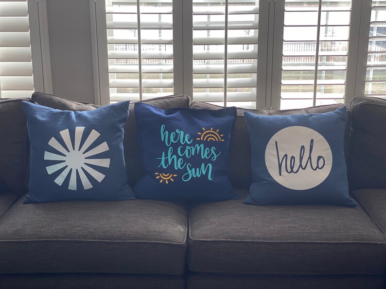 K&ouml;nnte beinhalten: Drei marineblaue Zierkissen mit wei&szlig;en Designs. Eines zeigt ein Sonnenstrahl-Design, ein anderes den Text "here comes the sun" mit Regenbogen und Sonne, und das dritte sagt "hello" in einem wei&szlig;en Kreis.