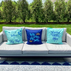 K&ouml;nnte beinhalten: Drei dekorative Kissen auf einem grauen Outdoor-Sofa. Die Kissen sind hellblau und marineblau, mit Texten und Grafiken wie "Life is better in Flip Flops", "Here comes the sun" und "Yes way Vacay!"