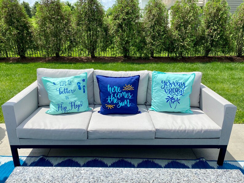 K&ouml;nnte beinhalten: Drei dekorative Kissen auf einem grauen Outdoor-Sofa. Die Kissen sind hellblau und marineblau, mit Texten und Grafiken wie "Life is better in Flip Flops", "Here comes the sun" und "Yes way Vacay!"