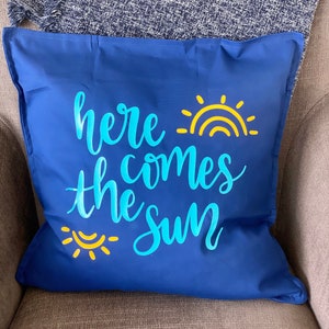 K&ouml;nnte beinhalten: Ein marineblaues quadratisches Kissen mit dem Schriftzug "here comes the sun" in hellblauer Schrift. Gelbe Sonne und Regenbogen-Grafiken akzentuieren den Text. Das Kissen liegt auf einem grauen Stuhl.