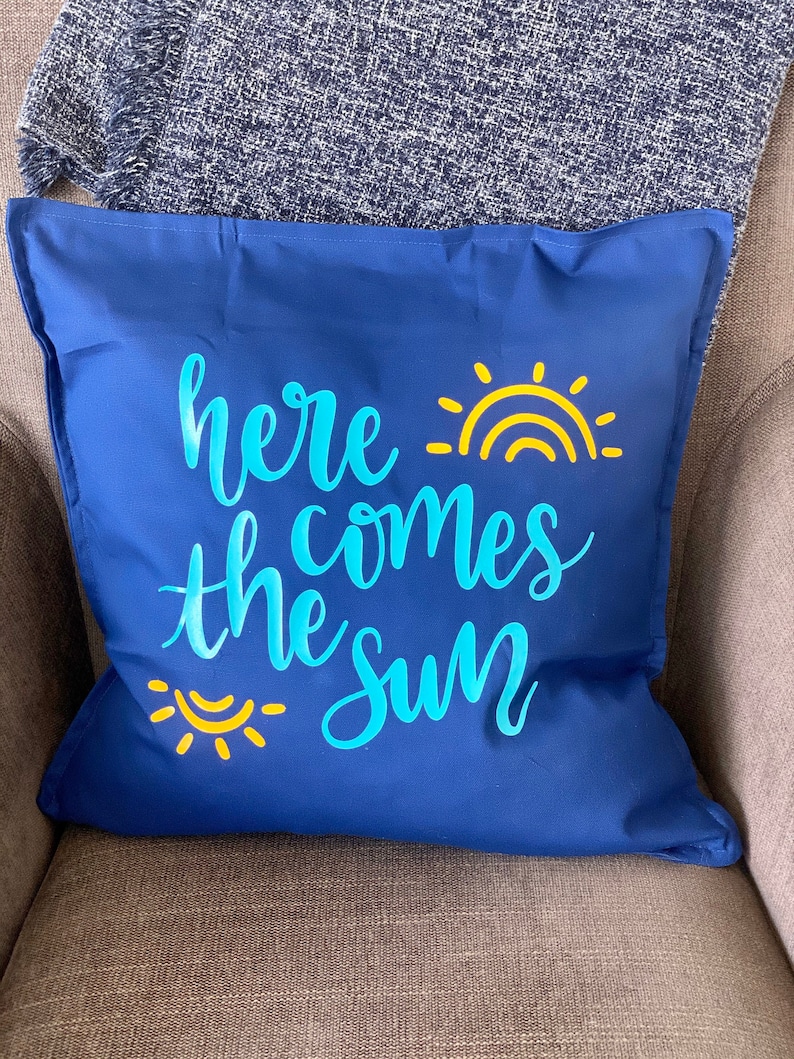 K&ouml;nnte beinhalten: Ein marineblaues quadratisches Kissen mit dem Schriftzug "here comes the sun" in hellblauer Schrift. Gelbe Sonne und Regenbogen-Grafiken akzentuieren den Text. Das Kissen liegt auf einem grauen Stuhl.