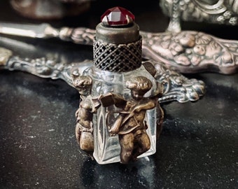 May include: A small glass bottle with a red glass stopper and two cherub figures on the sides. The bottle is clear and has a textured surface.