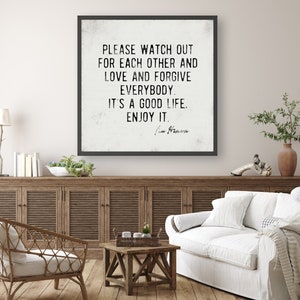 Op de afbeelding: Een ingelijste wandkunstprint met een zwart frame en een witte achtergrond. De tekst luidt: "PLEASE WATCH OUT FOR EACH OTHER AND LOVE AND FORGIVE EVERYBODY. IT'S A GOOD LIFE. ENJOY IT." De print wordt in een woonkamer getoond.