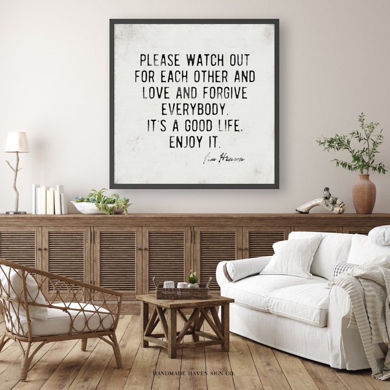Op de afbeelding: Een ingelijste wandkunstprint met een zwart frame en een witte achtergrond. De tekst luidt: "PLEASE WATCH OUT FOR EACH OTHER AND LOVE AND FORGIVE EVERYBODY. IT'S A GOOD LIFE. ENJOY IT." De print wordt in een woonkamer getoond.