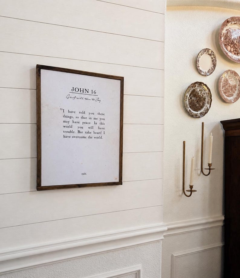 Puede incluir: Un marco de madera oscura con una impresi&oacute;n que dice "JOHN 16" y una cita sobre la paz. El marco est&aacute; colgado en una pared blanca con tablones. Tambi&eacute;n se ven platos decorativos y candelabros.