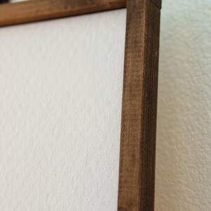 Puede incluir: Primer plano de un lienzo enmarcado. El marco es de madera marr&oacute;n oscuro, con vetas visibles y forma rectangular. El lienzo es blanco y tiene una superficie texturizada. Se ve la esquina del marco, mostrando la uni&oacute;n.