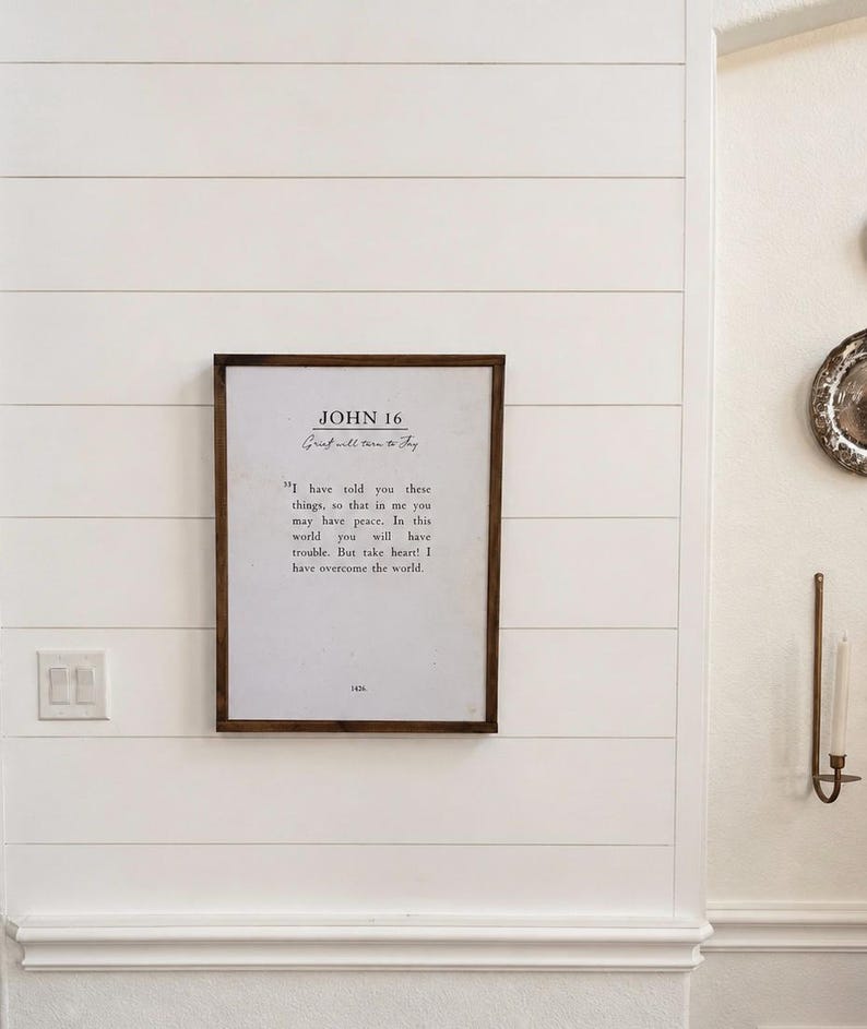 Puede incluir: Un marco de madera oscura con una impresi&oacute;n que contiene el texto &laquo;JOHN 16&raquo; y un vers&iacute;culo b&iacute;blico, colgado en una pared blanca con revestimiento de tablillas. El texto es negro. La impresi&oacute;n est&aacute; centrada en la pared.