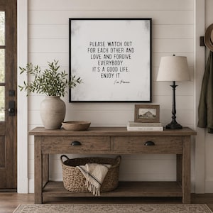 Op de afbeelding: Een rustieke houten console met twee lades en een onderste plank, versierd met een ingelijste quote, een lamp, een vaas met groen en een gevlochten mand. De ingelijste quote luidt: "PLEASE WATCH OUT FOR EACH OTHER AND LOVE AND FORGIVE EVERYBODY. IT'S A GOOD LIFE. ENJOY IT."