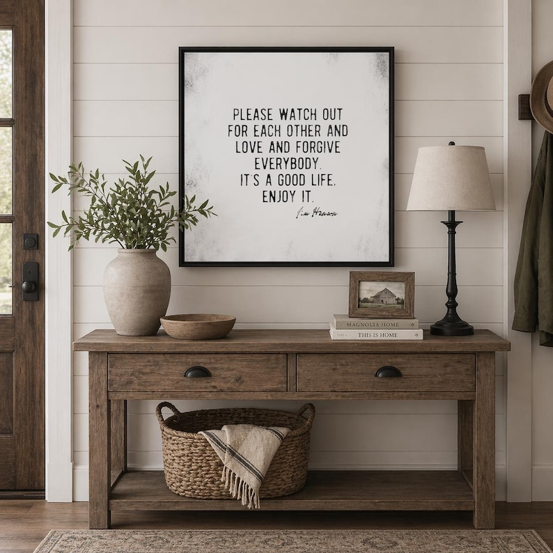 Op de afbeelding: Een rustieke houten console met twee lades en een onderste plank, versierd met een ingelijste quote, een lamp, een vaas met groen en een gevlochten mand. De ingelijste quote luidt: "PLEASE WATCH OUT FOR EACH OTHER AND LOVE AND FORGIVE EVERYBODY. IT'S A GOOD LIFE. ENJOY IT."