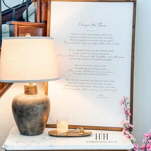以下が含まれることがあります： 「Prayer for Home」というテキストがエレガントな筆記体で書かれた額入りのサインが、大理石の天板が付いた木製のキャビネットに飾られています。ベージュのシェードと装飾的なベースのランプが隣にあり、キャンドルと花も添えられています。