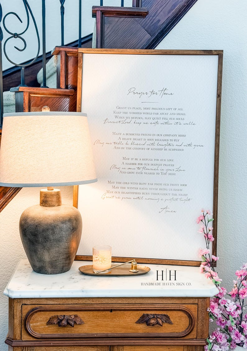 以下が含まれることがあります： 「Prayer for Home」というテキストがエレガントな筆記体で書かれた額入りのサインが、大理石の天板が付いた木製のキャビネットに飾られています。ベージュのシェードと装飾的なベースのランプが隣にあり、キャンドルと花も添えられています。