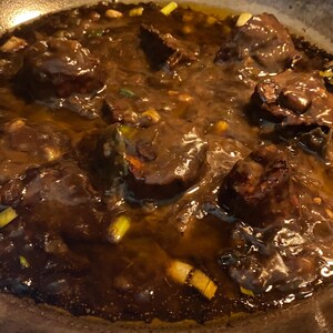 May include: Close-up of a pan filled with a dark brown stew. Large chunks of meat are visible, surrounded by a thick, glossy sauce. Green onions are scattered throughout the dish, adding a touch of color and freshness. The food is in a metal pan.