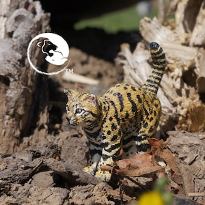 Puede incluir: Una pequeña figura realista de un ocelote, un gato salvaje con un pelaje manchado, de pie sobre un lecho de tierra marrón y hojas. El ocelote está mirando hacia la izquierda de la imagen.