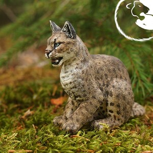 May include: A realistic figurine of a brown and white lynx sitting on a bed of green moss. The lynx has a spotted coat and is looking to the left.