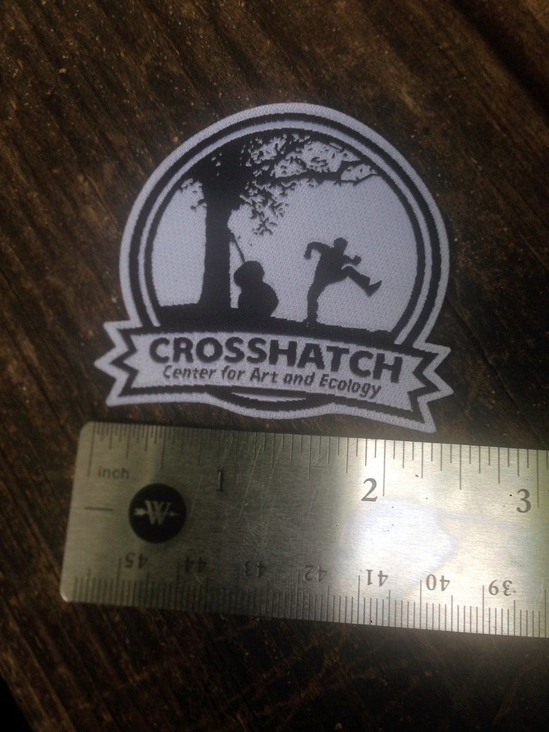 May include: White patch with black silhouette of a tree, a person running, and a cello. The patch has the text "CROSSHATCH Center for Art and Ecology". The patch is measured with a ruler, showing it is about 2 inches wide.