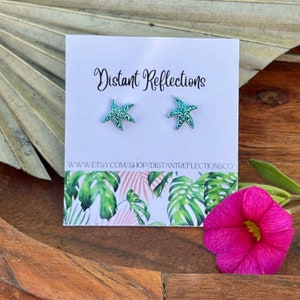 May include: A pair of teal blue glitter starfish stud earrings on a white card with the text "Distant Reflections" and the website address "www.etsy.com/shop/distantreflectionsco".