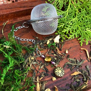 May include: A silver tea infuser ball with a chain and a decorative charm. The charm is a gold-colored flower with a beaded chain attached. The infuser is sitting on a wooden surface with green moss and loose tea leaves.