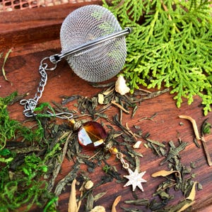 May include: A silver tea infuser with a chain and a decorative amber crystal bead and star charm. The infuser is sitting on a bed of loose leaf green tea.