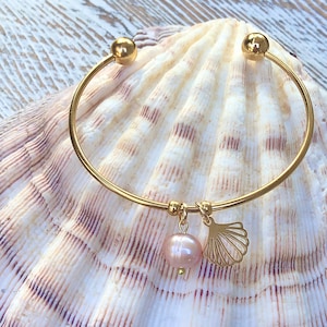 May include: Gold-toned bangle bracelet with a pink pearl and a seashell charm. The bracelet has a simple, elegant design with a round pearl and a delicate seashell pendant. The bracelet is displayed on a large seashell.