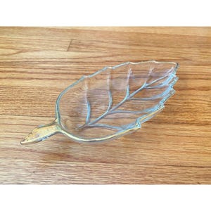 May include: A clear glass leaf-shaped dish with gold trim. The dish has a detailed vein pattern and is set against a wooden surface. This decorative dish is ideal for serving snacks or as a decorative accent.
