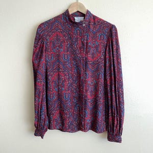Pode incluir: Uma blusa de manga comprida com gola alta e fecho frontal de botões. A peça é decorada com um padrão paisley denso e complexo em tons de vermelho profundo, azul marinho e turquesa brilhante, com punhos franzidos nas mangas.