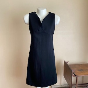 May include: A sleeveless, knee-length black dress displayed on a mannequin. The dress features a V-neckline and a fitted silhouette. The fabric appears smooth and solid, with a subtle sheen. The dress is a classic, versatile garment.