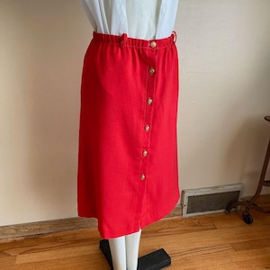 May include: A vibrant red skirt featuring a button-up design with gold-toned buttons. The skirt has an elasticated waistband and falls to knee length. The fabric is a solid colour.