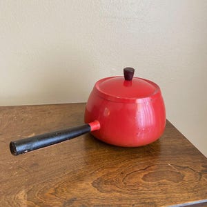 Może przedstawiać: Jasnoczerwony garnek do fondue z pasującą pokrywką i długą, prostą czarną rączką. Pokrywka posiada małe, ciemnobrązowe pokrętło na środku. Garnek ma zaokrąglony, pękaty kształt oraz gładkie, błyszczące wykończenie na całej zewnętrznej powierzchni.