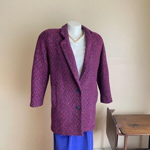 May include: A magenta and black herringbone pattern coat with a notched collar and two dark blue buttons. The coat is displayed on a mannequin with a white top and a gold necklace. The bottom half of the mannequin is wearing a blue skirt.