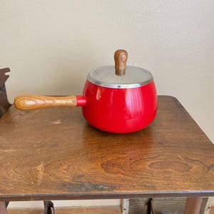 Może przedstawiać: Mały, czerwony rondel z drewnianym uchwytem i pokrywką. Rondel ma srebrną obręcz i drewnianą gałkę na pokrywce. Uchwyt jest również wykonany z drewna. Rondel stoi na drewnianym stole.