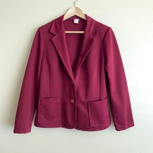Puede incluir: Un blazer burdeos con cierre de un solo botón y dos bolsillos delanteros. La chaqueta está colgada de una percha de madera sobre un fondo neutro. El blazer tiene un diseño clásico y entallado.