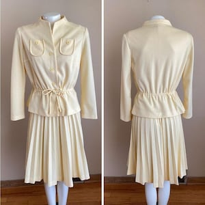 May include: A vintage pale yellow two-piece suit. The jacket has a mandarin collar, two front pockets, and button closures. The skirt is pleated. The suit is displayed on a mannequin.