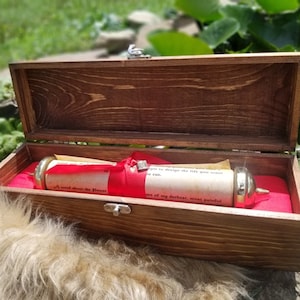 May include: A wooden box with a red velvet lining, containing a rolled up scroll with a red ribbon and gold accents. The scroll has text on it that reads "A word about the Power... to design the life you want... some of my darkest, most painful..."