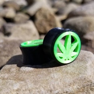 Puede incluir: Tapones para los oídos negros y verdes con un diseño de hoja de cannabis. Los tapones están hechos de un material oscuro con un recorte de hoja verde brillante. Los tapones están sobre rocas.