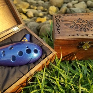 May include: A blue ocarina with a glittery finish rests in an open wooden box lined with black fabric. A second wooden box with a carved design is next to it. The ocarina has a triangular emblem.