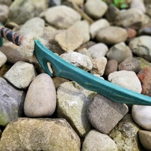 May include: A teal-colored fantasy sword with a textured blade and a green handle wrapped in black and brown material. The sword is resting on a bed of various-sized rocks, creating a natural, outdoor setting.
