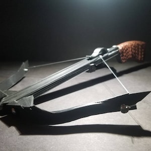 May include: A black crossbow with a brown grip and a white string is set against a dark background. The crossbow is made of black material and has a detailed design. The image is well-lit, highlighting the crossbow's features.