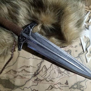 Puede incluir: Una daga de plata con mango de cuero marrón y una intrincada metalurgia en la empuñadura. La daga está sobre un mapa con un recorte de dragón blanco encima.