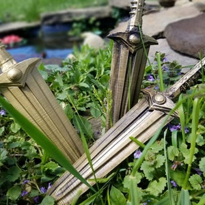 May include: Three gold-colored daggers with intricate designs are displayed in a bed of green foliage with purple flowers. The daggers have a sharp point and a textured handle.