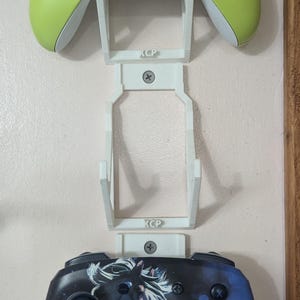 May include: A wall-mounted gaming controller holder with two controllers. The top controller is lime green, and the bottom controller has a custom design. The holder is white and has the letters "KCP" printed on it.