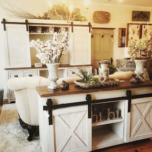 May include: White farmhouse style cabinet with sliding barn doors. The cabinet has a wood top and a white base with black hardware. The cabinet has two shelves and a bottom drawer. The cabinet is decorated with a vase of cotton flowers and a tray of greenery.