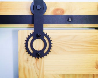 May include: Black metal sliding door hardware with a gear-shaped design. The hardware is mounted to a light wood door.
