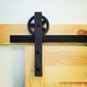 May include: Black metal sliding barn door hardware with a round wheel and a black track. The hardware is mounted on a light wood door.