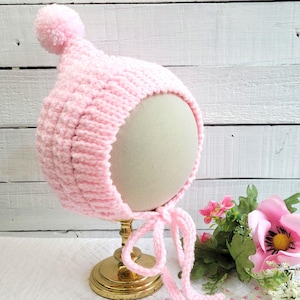 May include: A pink crocheted baby bonnet with a pom-pom on top and ties at the bottom.