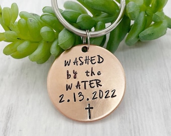 May include: A copper-coloured keychain with a round tag that reads "WASHED by the WATER 2.13.2022" with a small cross below the date. The keychain is attached to a silver key ring.