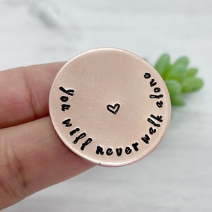 May include: A rose gold coloured metal disc with the words "You will never walk alone" engraved around the edge. A small heart is engraved in the centre of the disc.