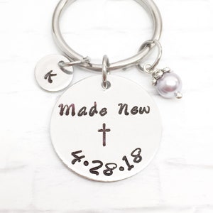 May include: Silver keychain with a round charm that says "Made New" with a cross and the date "4.28.18". The keychain also has a small round charm with the letter "K" and a pearl charm.