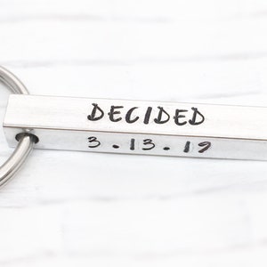 May include: Silver key chain with a rectangular bar engraved with the words "DECIDED" and the date "3.13.19".