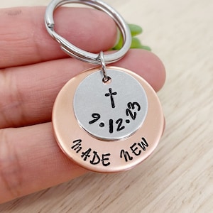 Op de afbeelding: Een sleutelhanger van zilver en koper met een kruis en de datum 9.12.23 aan de ene kant en de woorden "Made New" aan de andere kant.
